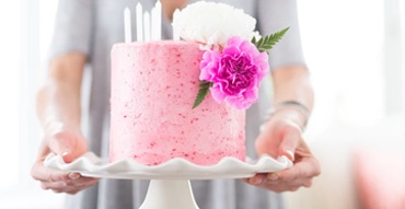 A fancy pink birthday cake with white candles and pink and white fresh flowers is on a white cake plate and hold by a lady.